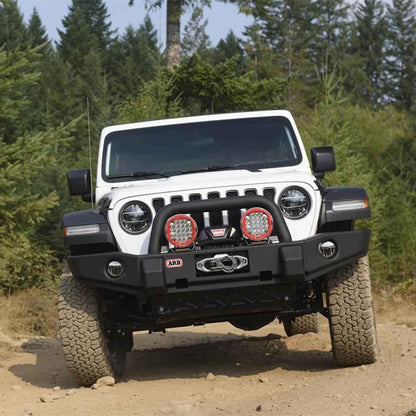 White Jeep Wrangler with additional lights on a dirt road surrounded by trees
