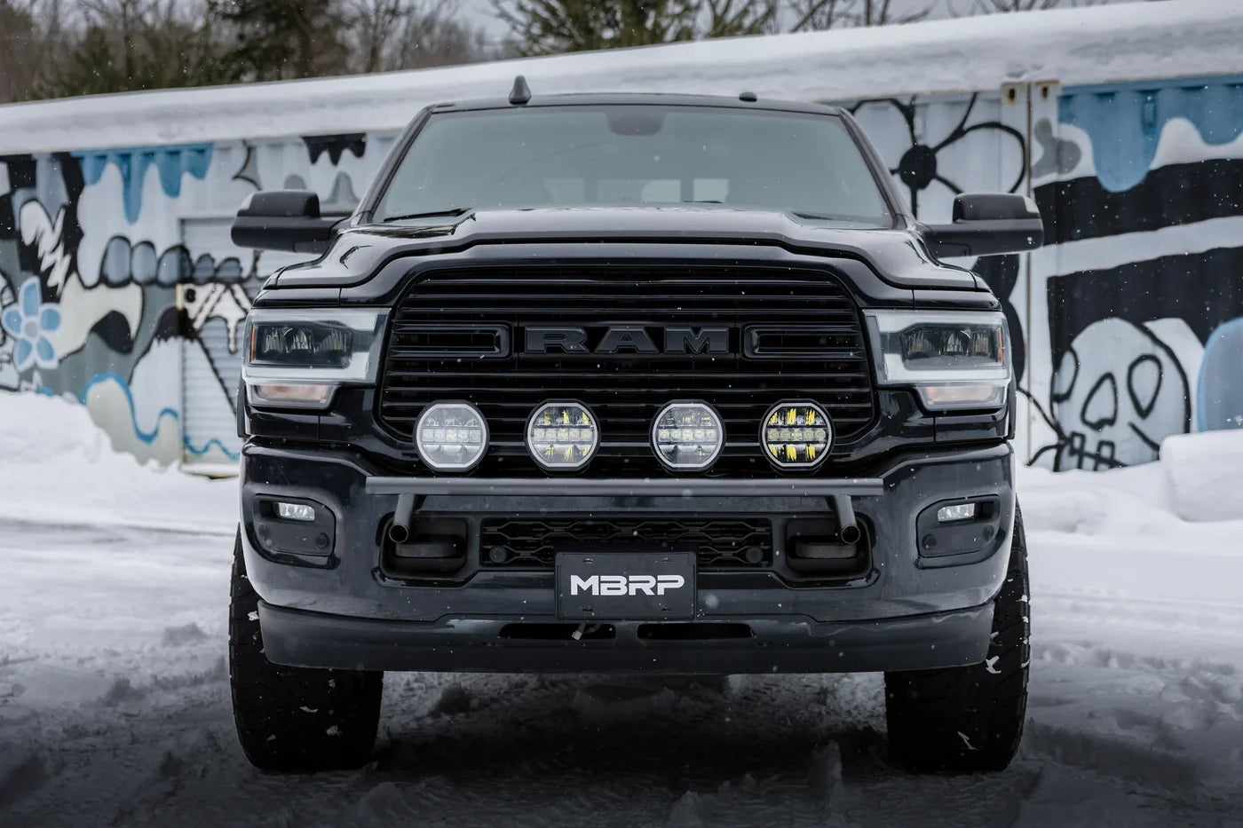 A black Ram pickup truck photographed head-on in a snowy outdoor setting with a graffiti-covered wall behind it. The front grille shows the Ram logo, and the lower bumper plate reads MBRP.