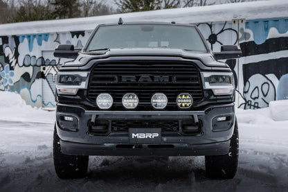 A black Ram pickup truck photographed head-on in a snowy outdoor setting with a graffiti-covered wall behind it. The front grille shows the Ram logo, and the lower bumper plate reads MBRP.